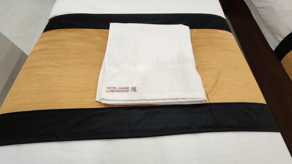 A neatly made bed with a white towel, black and brown bedspread, and a tag reading 'HOTEL GRAND LUSHWOOD.'