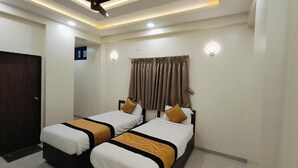 A hotel room with two beds, a ceiling fan, recessed lighting, a window with curtains, and a door. - Hotel Grand Lushwood (Chhatrapati Sambhajinagar)