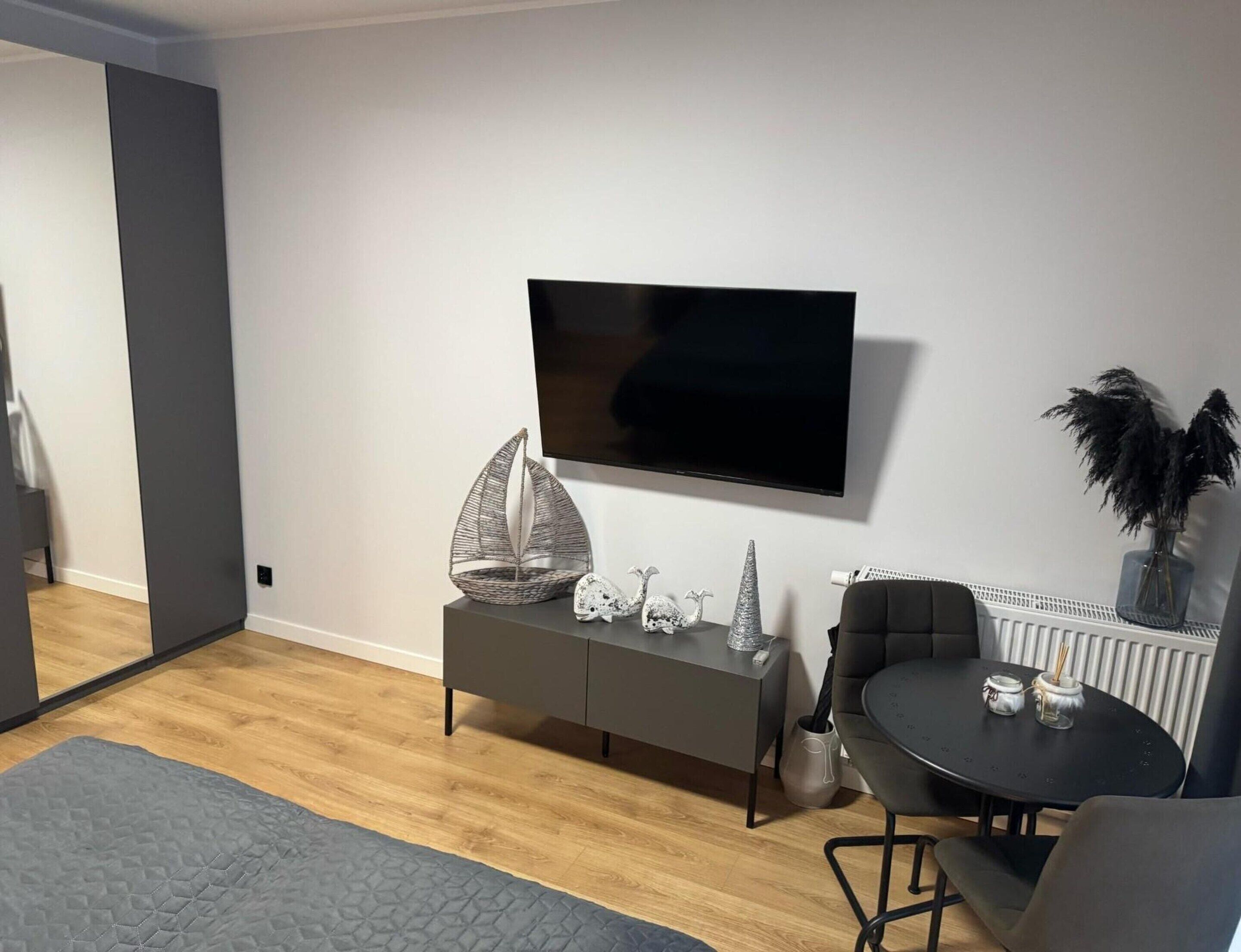 A modern living room with a flat-screen TV mounted on the wall, a grey sofa, a small round table with two chairs, and decorative items on the sofa.