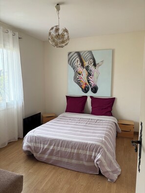 A bedroom with a bed, two pillows, a painting of zebras, a ceiling light fixture, and a window with curtains. - Charming renovated Landes house (Mimizan)