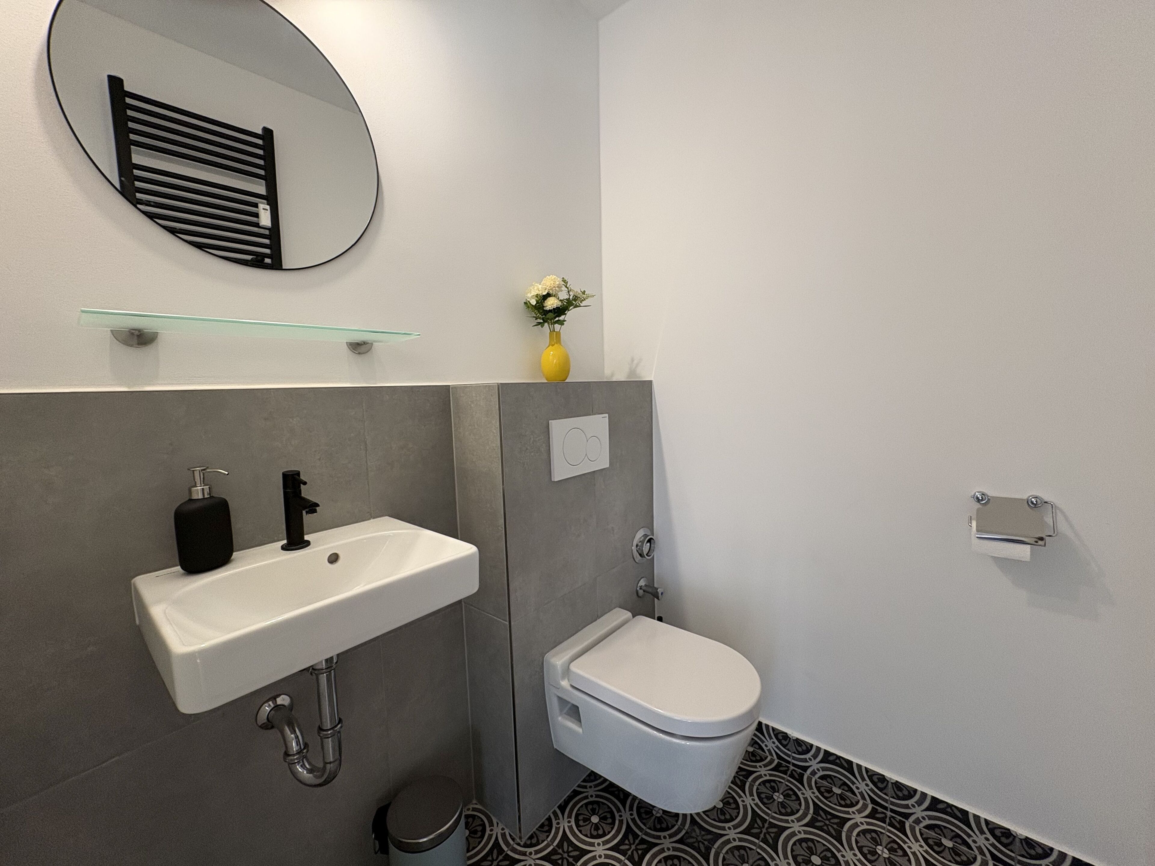 A bathroom with a white sink, black faucet, and a toilet. There is a mirror above the sink and a small vase with flowers on the wall.