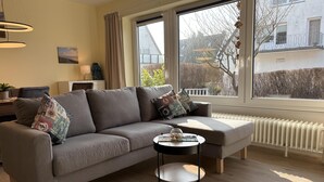Un salon moderne avec un canapé gris, des coussins à motifs, une table basse ronde et une grande fenêtre donnant sur un immeuble et des arbres.