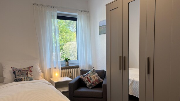 Une chambre à coucher comprenant un lit, une chaise, une armoire, une fenêtre avec des rideaux et un radiateur.