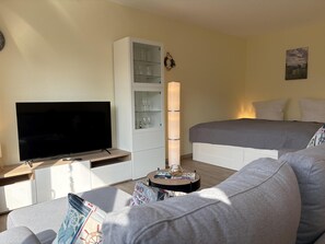 Ein Hotelzimmer mit Flachbildfernseher, einem weißen Schrank mit Glastüren, einem Nachttisch mit Lampe, einem grauen Sofa und einem Bett mit weißer Bettwäsche.