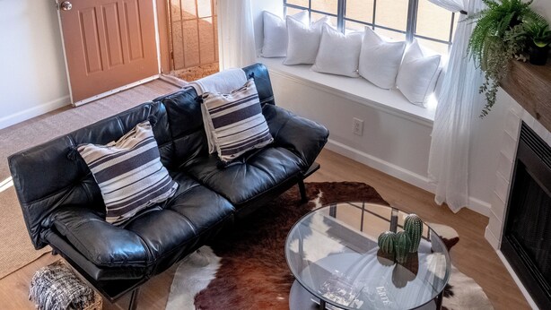 Una sala de estar con un sofá de cuero, una mesa de centro de vidrio, un tapete de piel de vaca, una lámpara colgante, una ventana con cortinas blancas y una puerta.