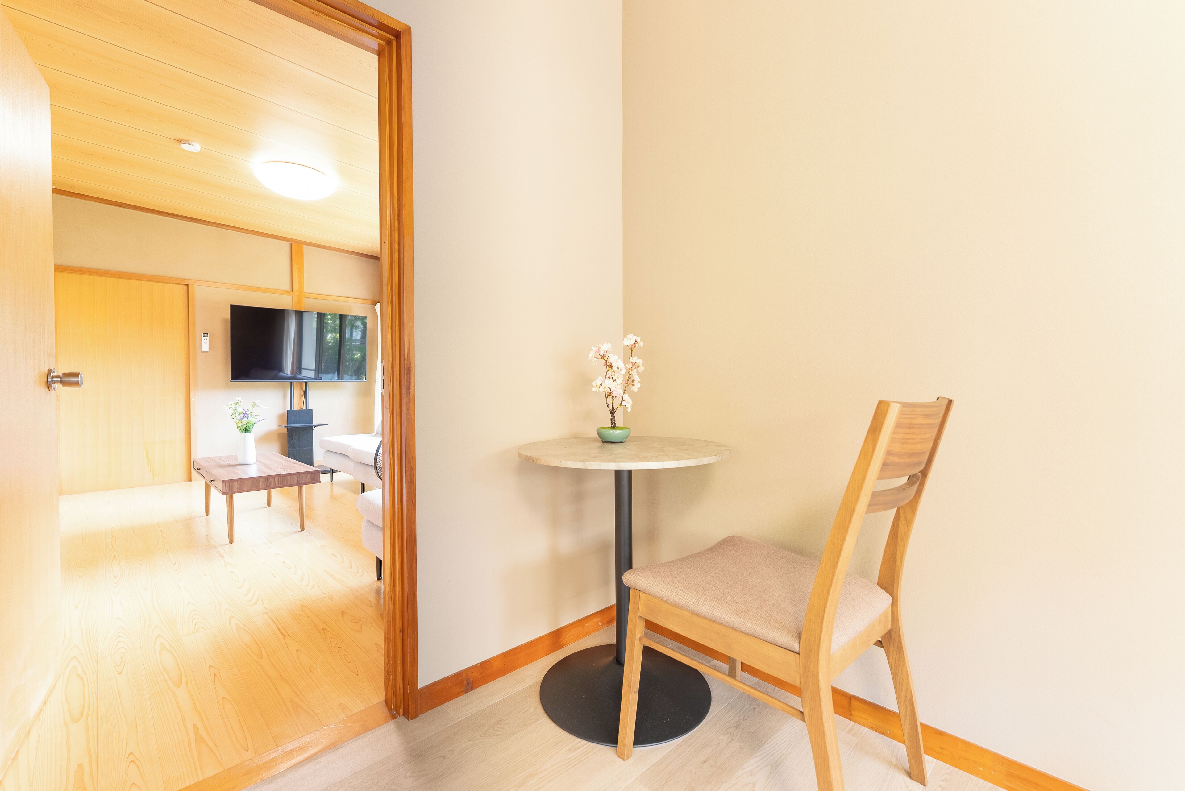 Une pièce avec un plancher en bois, une table ronde surmontée d’un vase, une chaise, un téléviseur accroché au mur et une porte donnant sur une autre pièce.