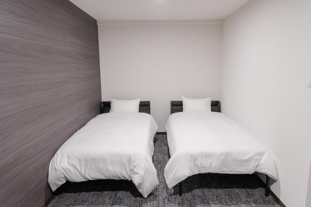 A small, modern room with two single beds, a grey textured wall, and a carpeted floor.