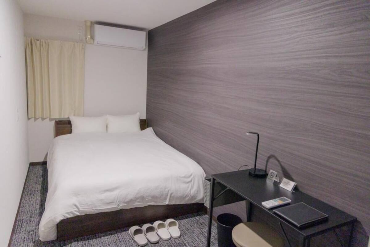 A hotel room with a bed, a desk, a chair, a trash bin, and a pair of slippers.