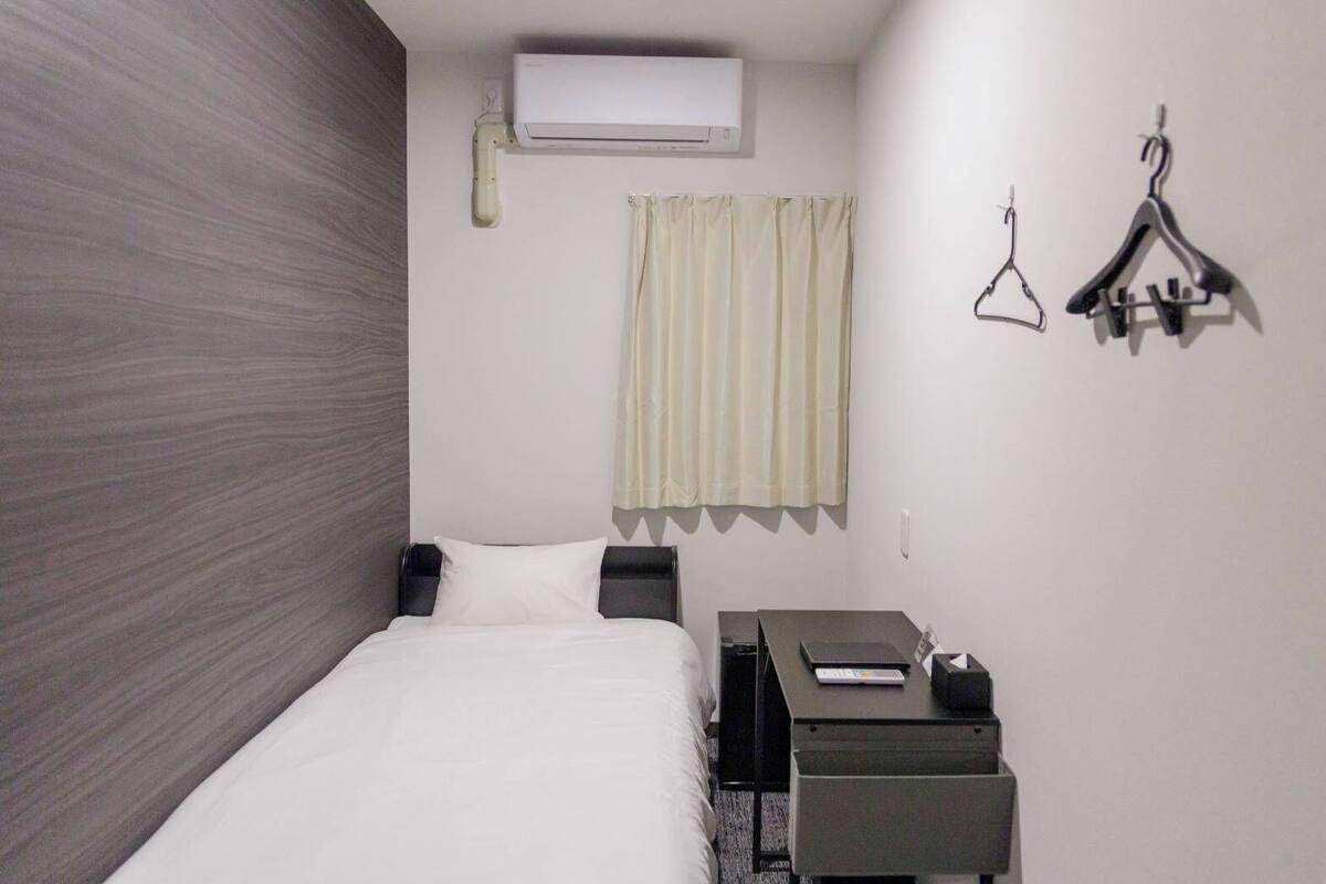 A hotel room with a bed, a desk with a computer, an air conditioner, a curtain, and two hangers on the wall.