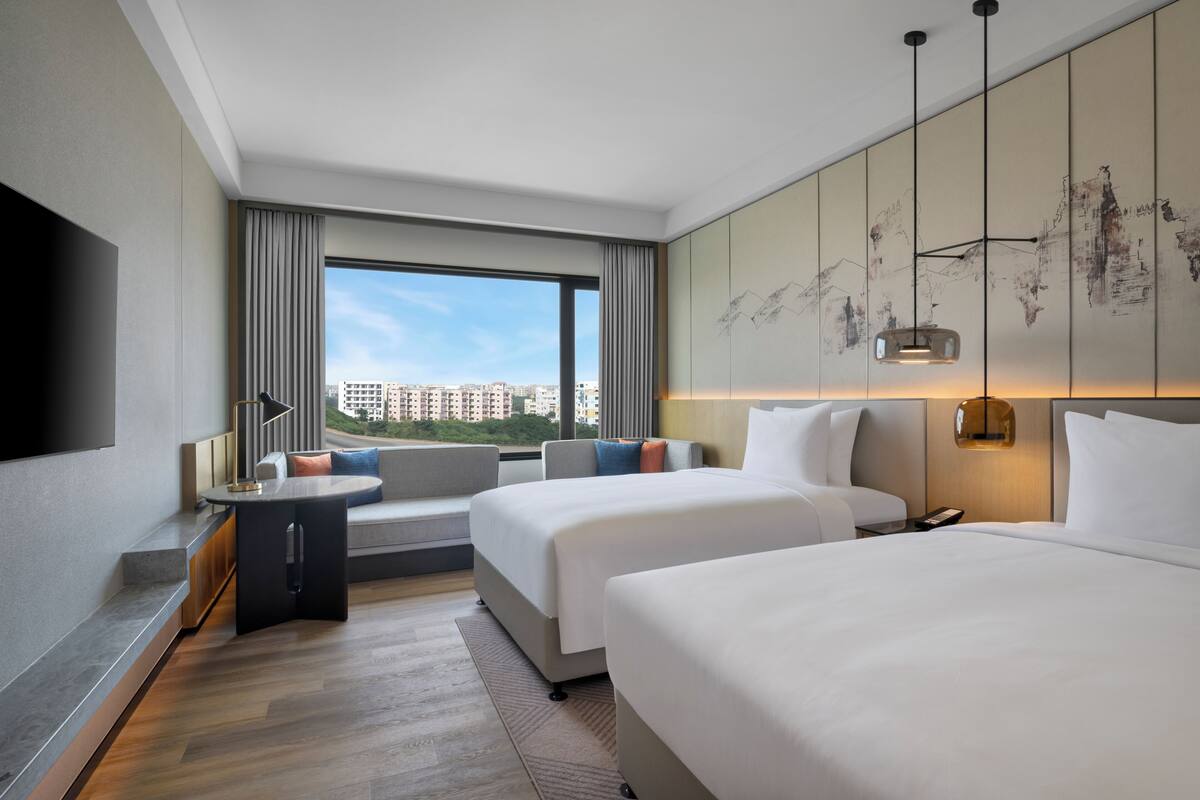 A hotel room with a large window, a sofa, two beds, a flat-screen TV, and a wall-mounted artwork.