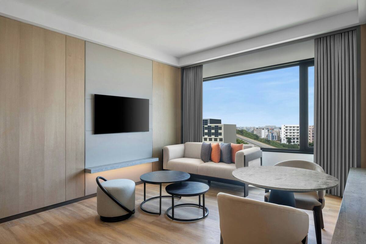 A modern hotel room with a sofa, two round tables, a flat-screen TV, and a large window with a city view.