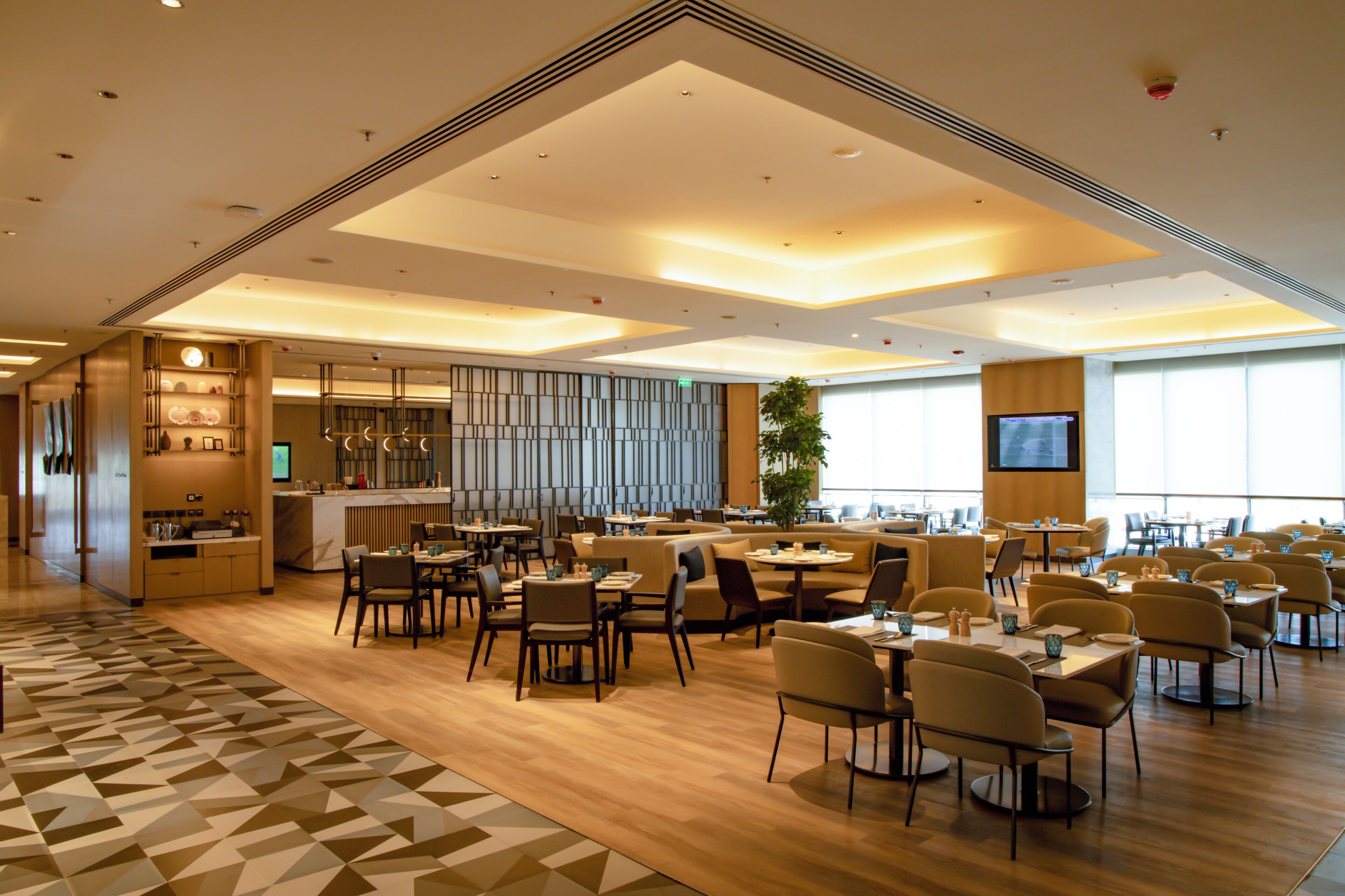 A spacious hotel lobby with a central bar, multiple seating areas, and a large screen for presentations.