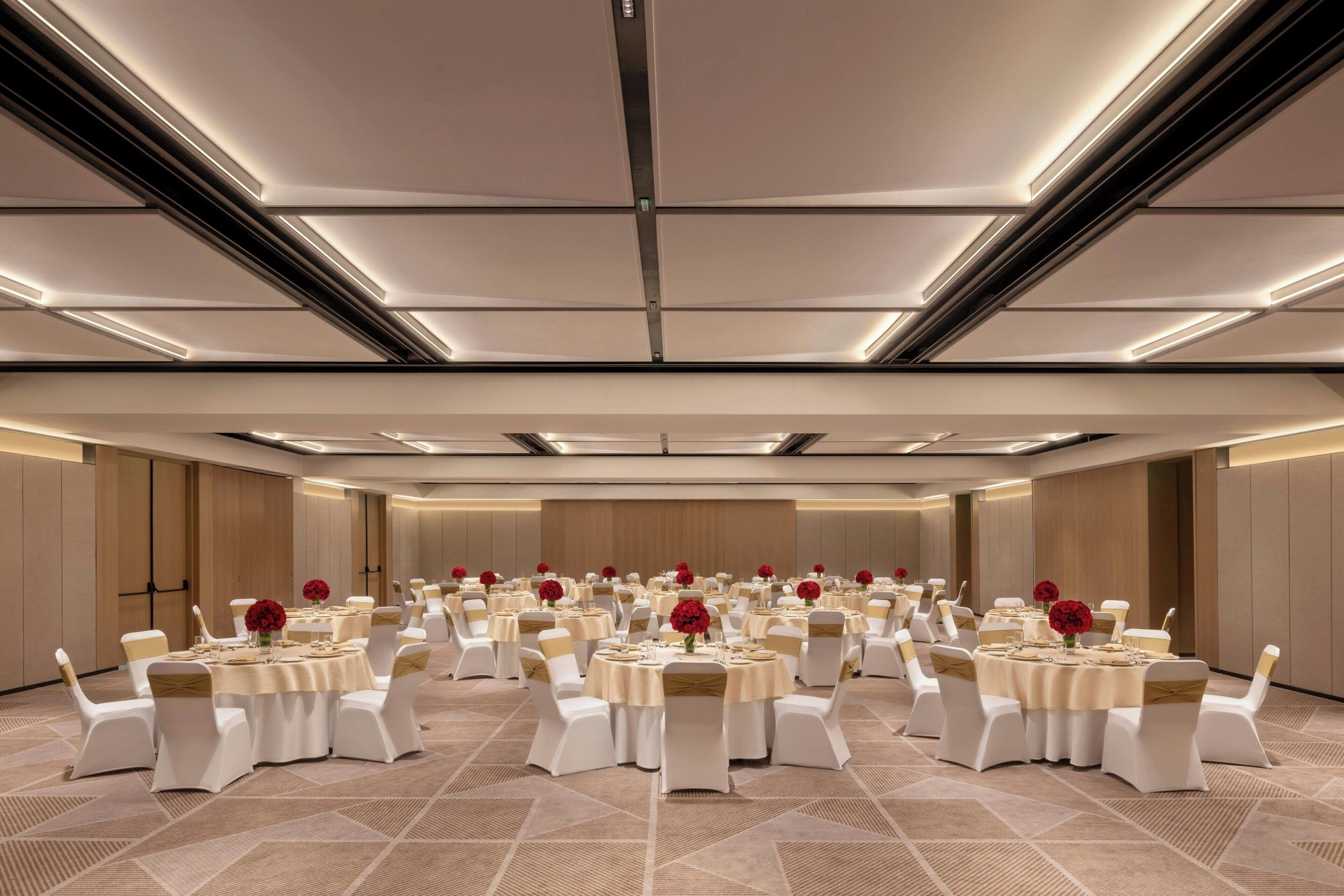 A large conference room with multiple round tables, each with white chairs and red flowers.