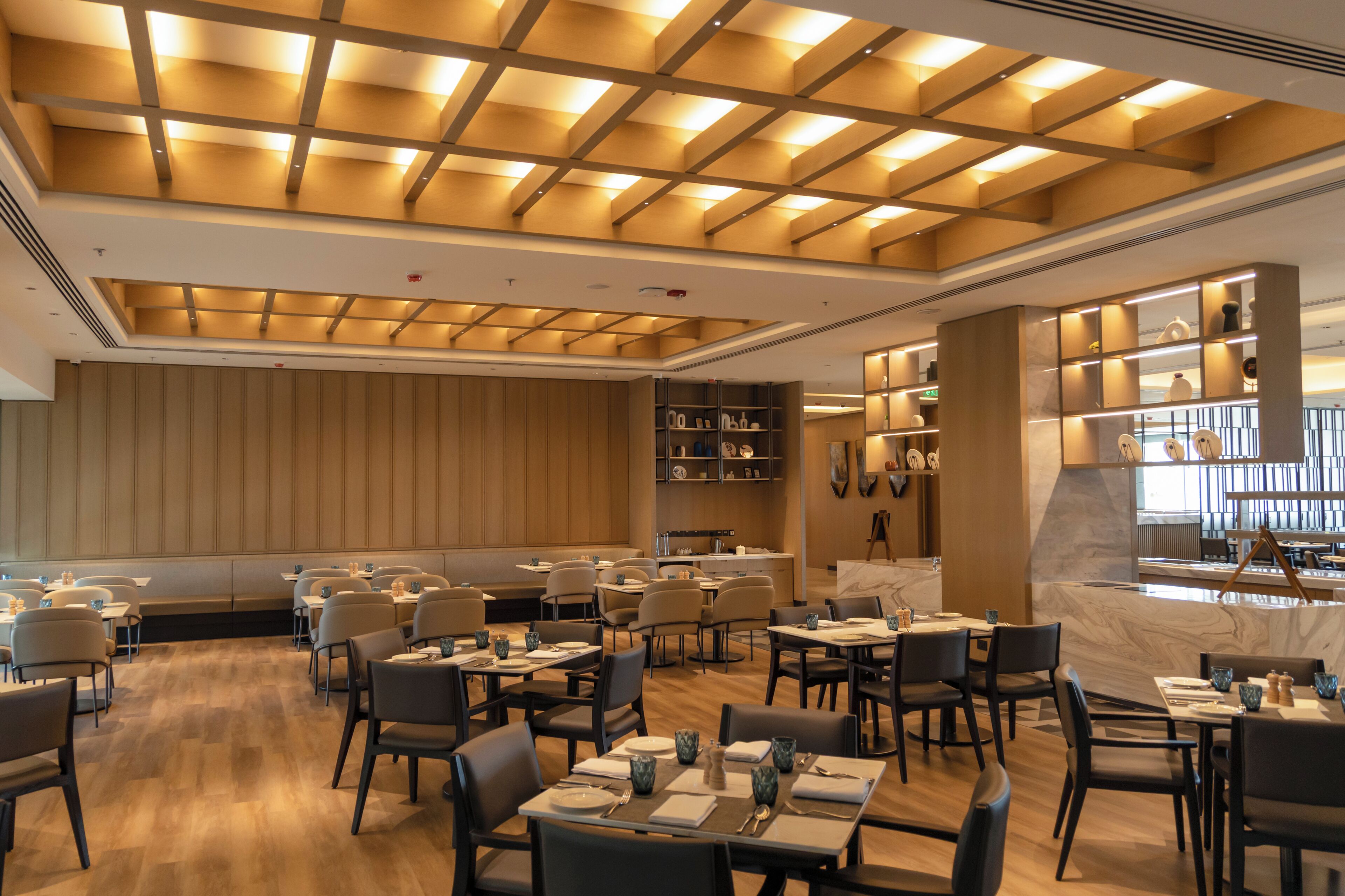 A modern restaurant with a warm ceiling design, wooden panel walls, and a marble counter. Tables are arranged for dining.