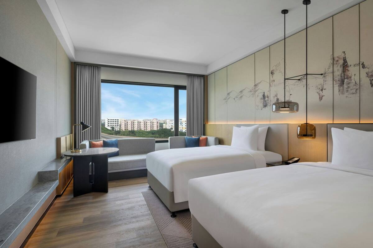 A hotel room with two beds, a sofa, a TV, and a large window with a city view.
