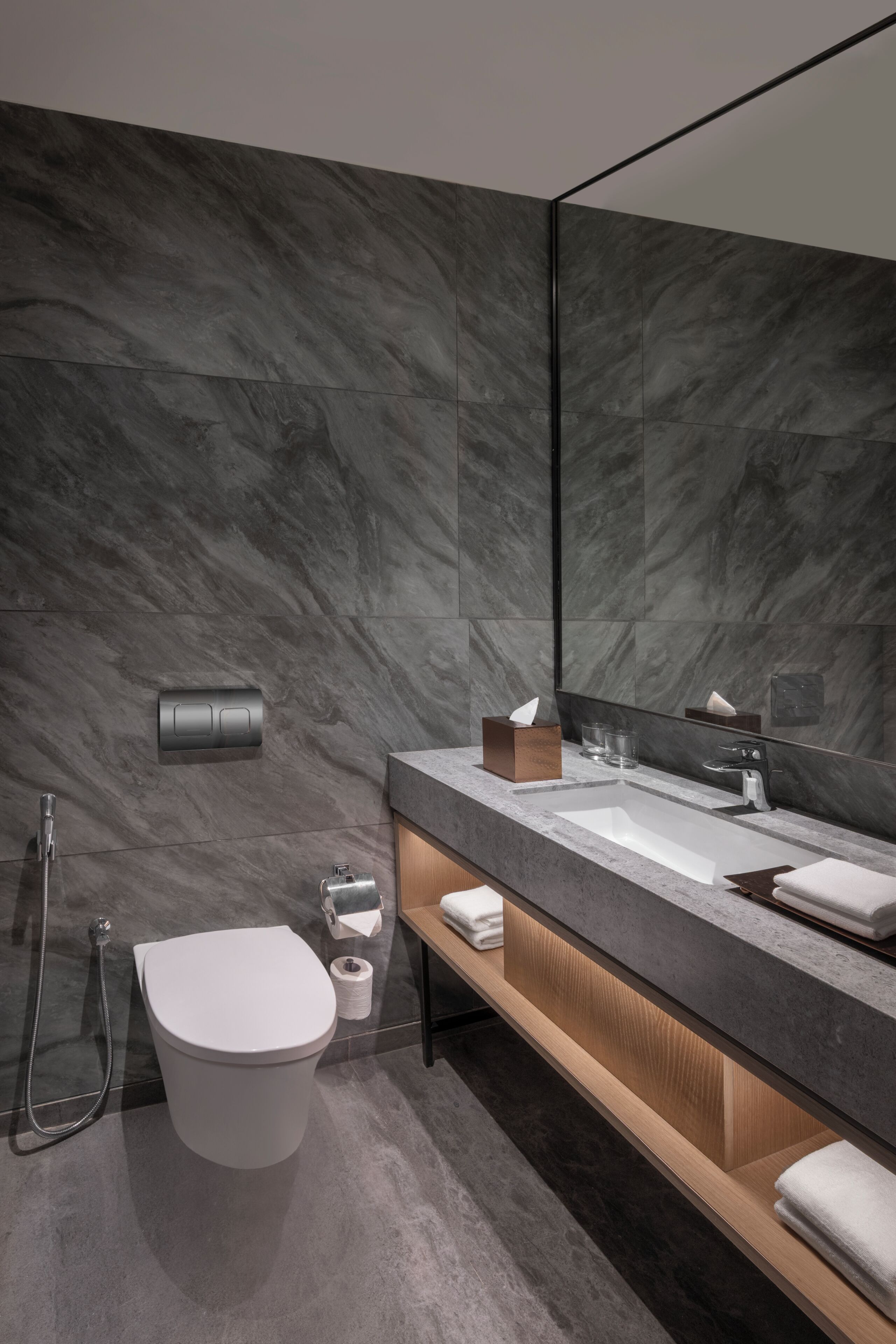 Een moderne badkamer met een marmeren wand, een grote spiegel, een wit toilet, een wastafel met houten kastje en een handdoekrek.