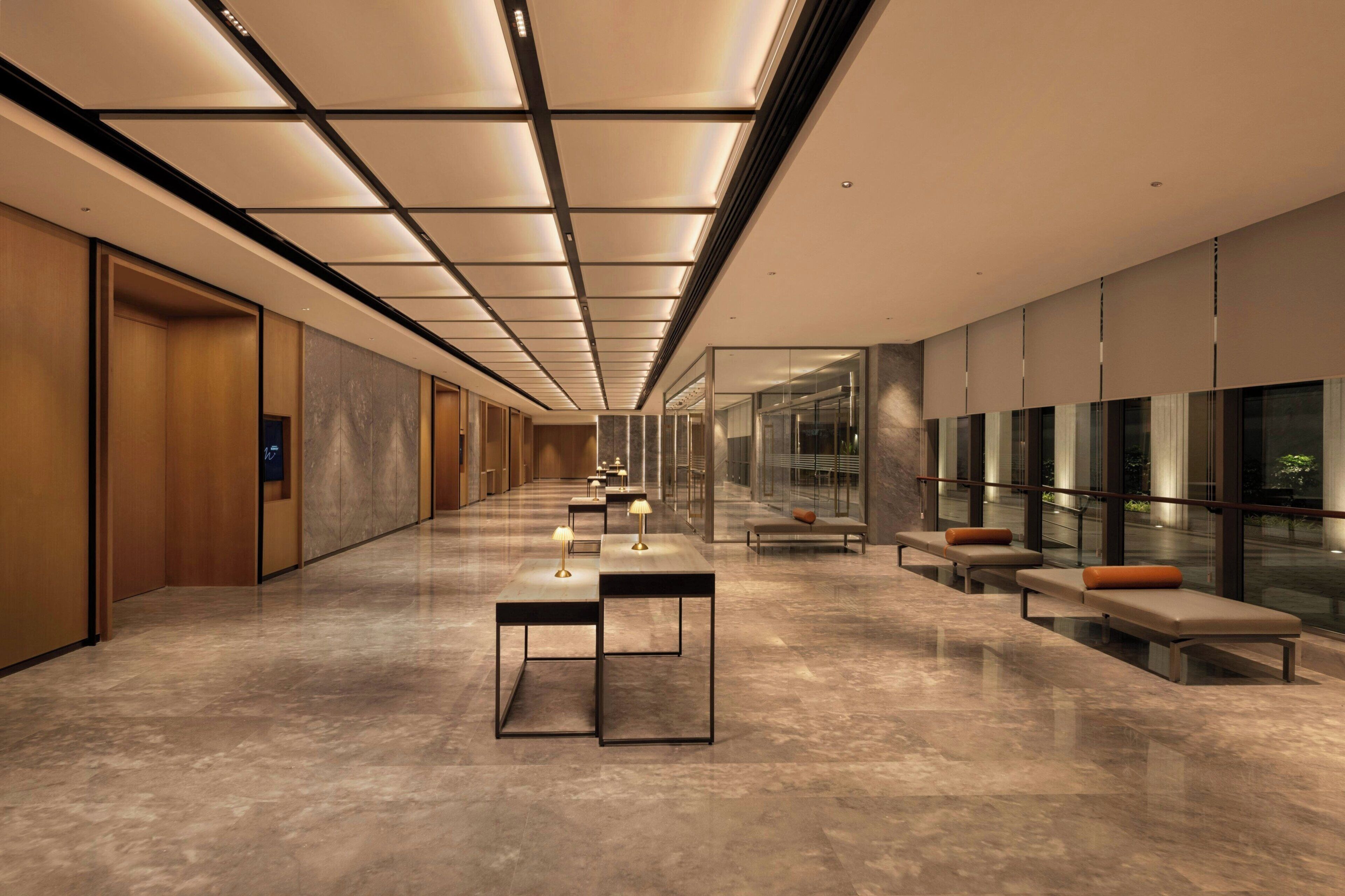 A modern hotel corridor with a long hallway, seating areas, and a glass wall on one side.