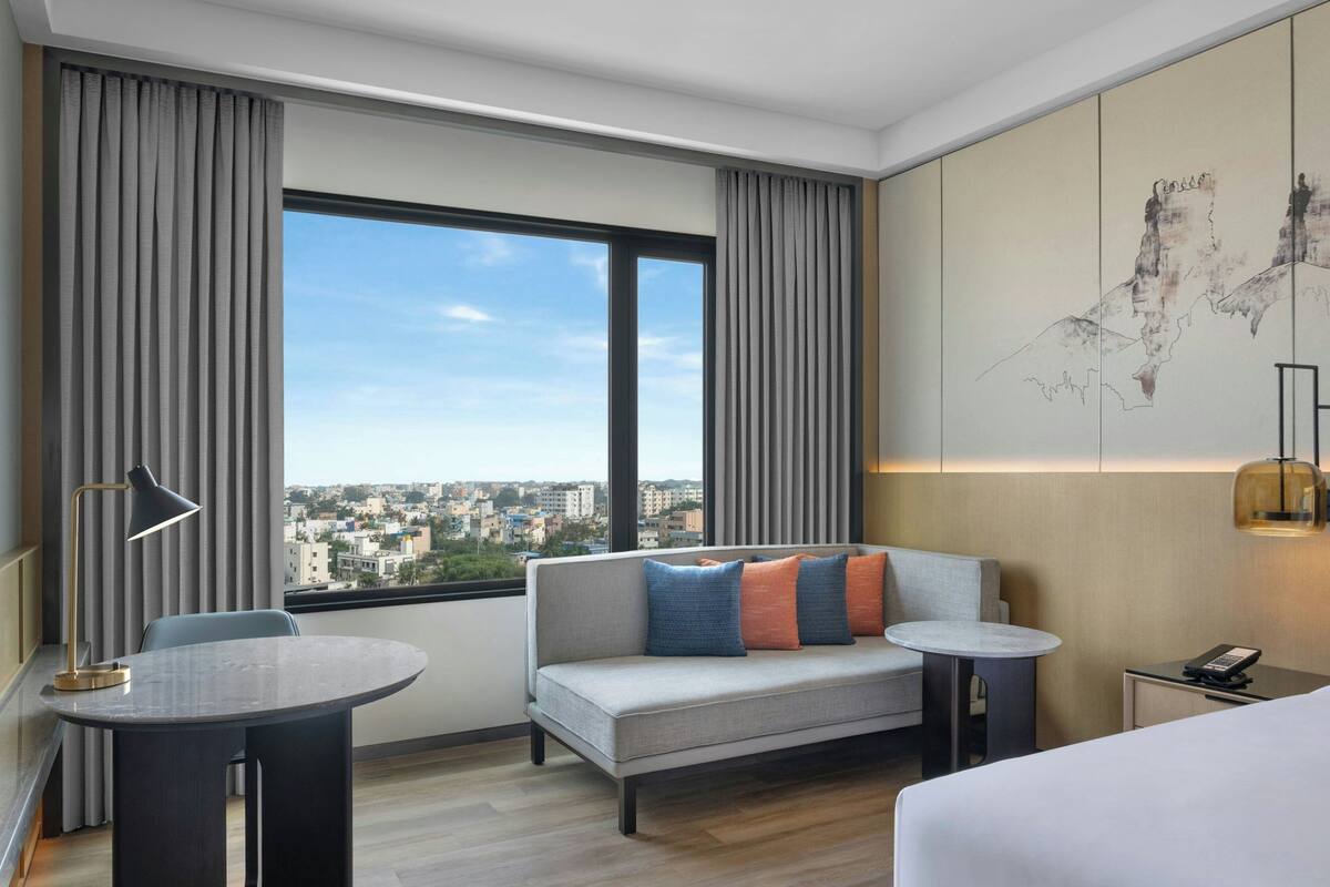 A modern hotel room with a sofa, a small table, a bed, a desk, and a view of a cityscape through large windows.