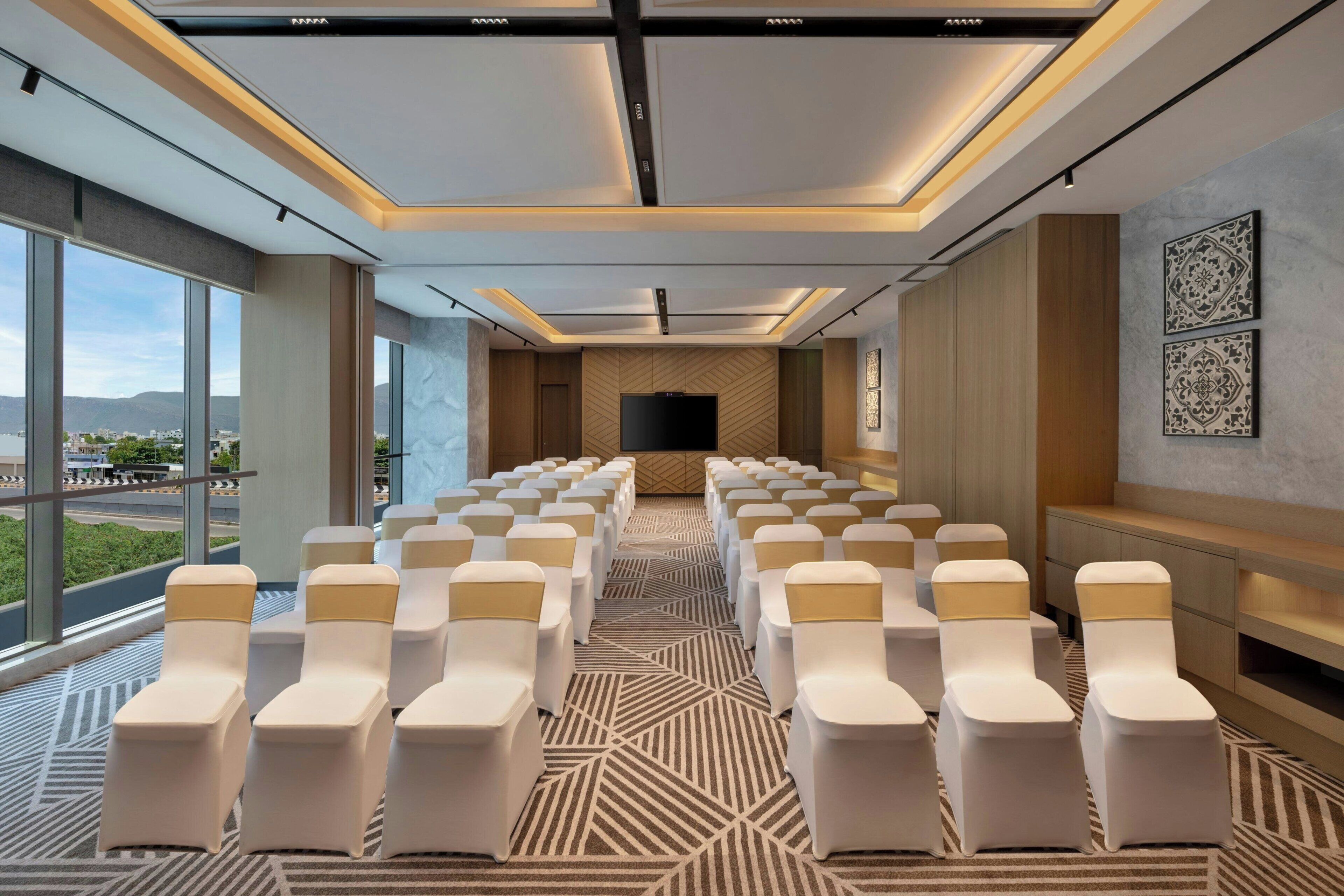 Un salón de conferencias con filas de sillas, una pantalla grande y vistas al exterior.