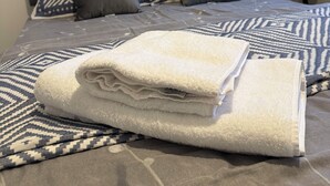 Una cama bien tendida con una colcha estampada, dos almohadas, una toalla blanca doblada y una mesita de noche con una tetera.