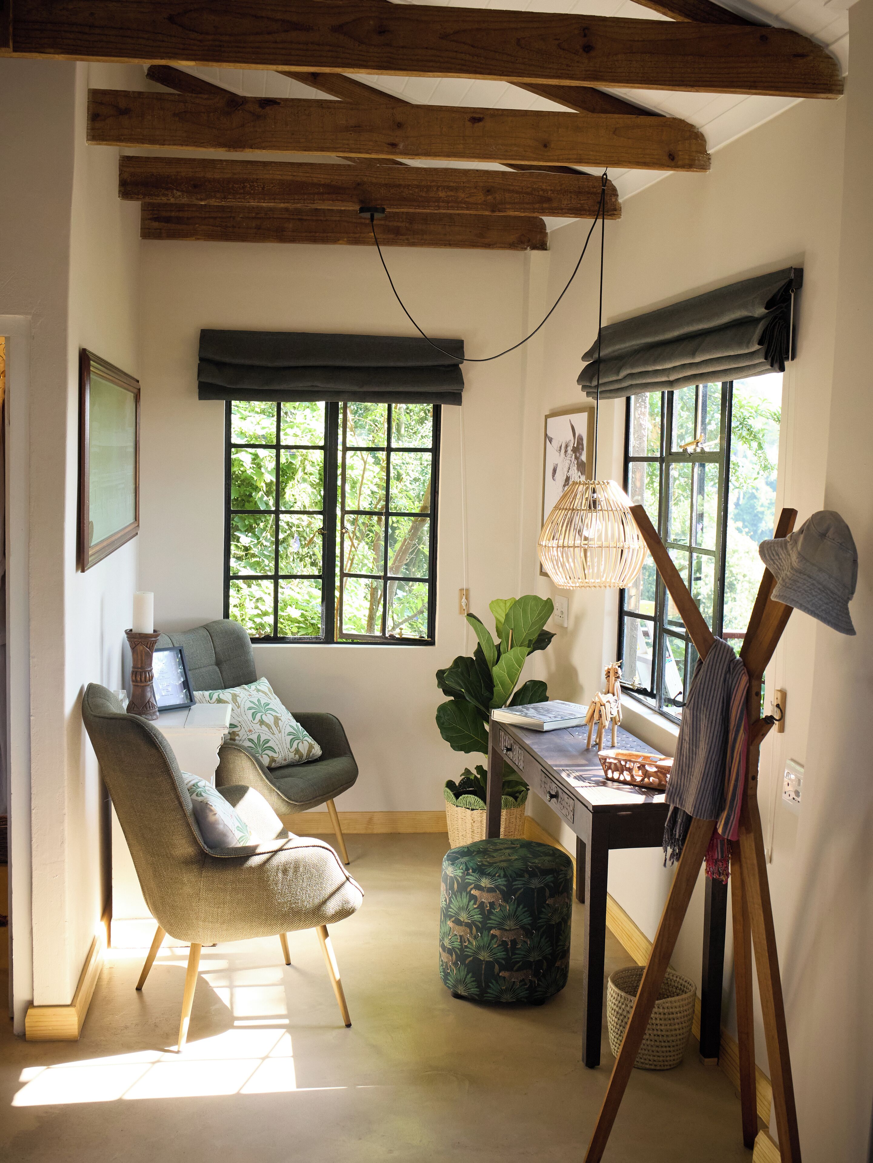 Una stanza con soffitto in legno, una finestra vista verde, una sedia, un tavolo con una lampada e un quadro appeso al muro.