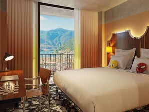 A hotel room with a bed, a desk with a chessboard, a chair, a lamp, and a view of mountains. - Mama Shelter Lake Como (Como)