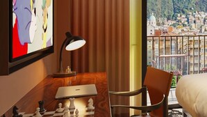 A hotel room with a wooden desk, a laptop, a chessboard, a lamp, a bed, and a large window with a city view. - Mama Shelter Lake Como (Como)