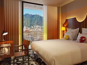A hotel room with a bed, a desk with a chessboard, a chair, a lamp, and a view of a cityscape. - Mama Shelter Lake Como (Como)