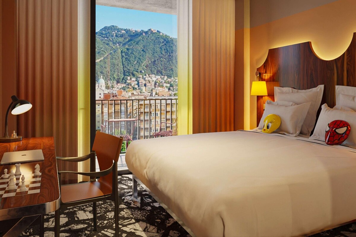 A hotel room with a bed, a desk with a chessboard, a chair, a lamp, and a view of a cityscape.