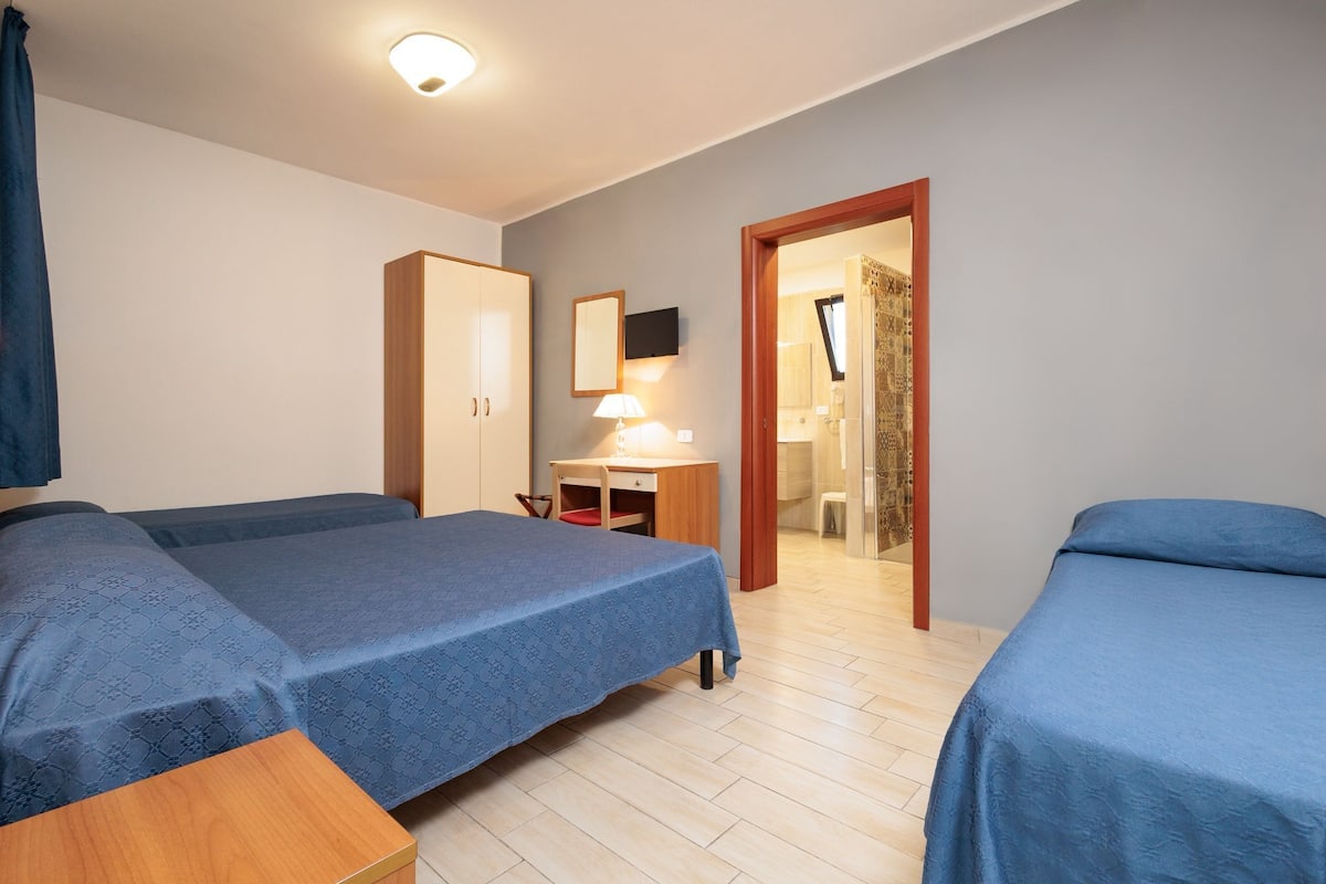 A bedroom with two beds, a desk, a wardrobe, and a bathroom visible through an open door.