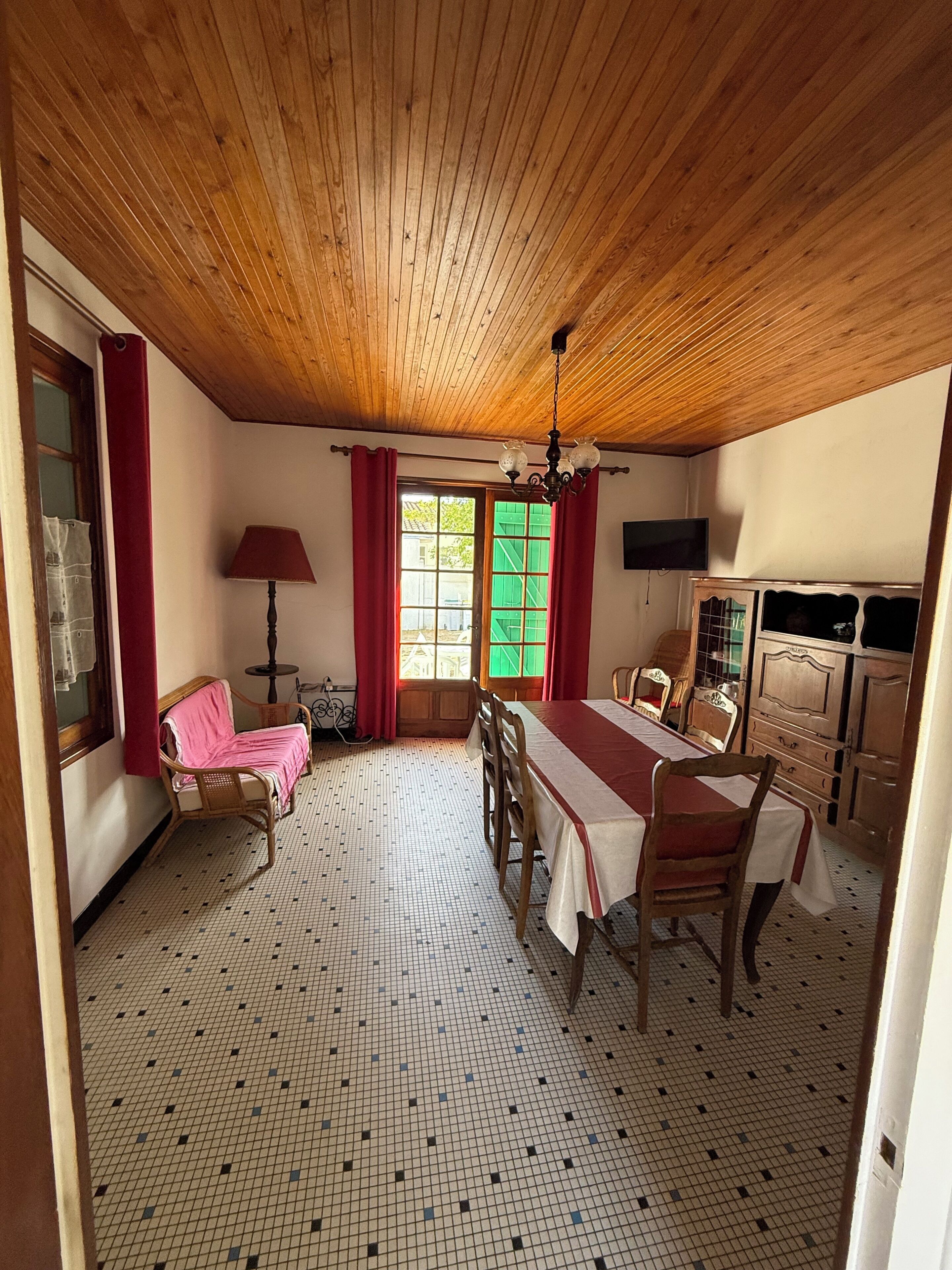 A room with a wooden ceiling, a dining table with chairs, a sofa, a TV, and a window with curtains.