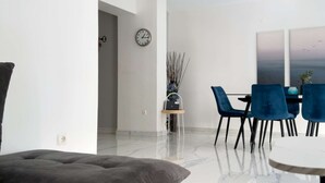 Ein modernes Wohnzimmer mit einer Couch, einem Essbereich mit blauen Stühlen, einer Uhr an der Wand und einer Vase mit Zweigen.