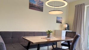 Een zithoek met een houten tafel, een grijze gestoffeerde bank en twee stoelen. Twee hanglampen, een potplant en een lamp op een bijzettafeltje.