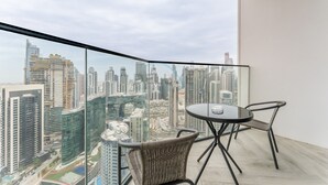 Un balcón con una barandilla de vidrio, una mesa redonda, dos sillas de mimbre y vista panorámica de la ciudad.
