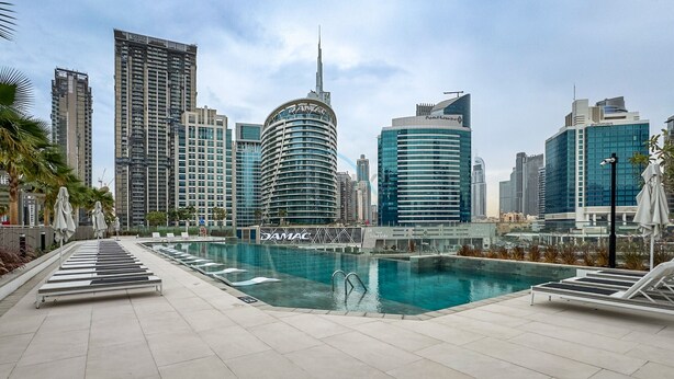 Un área de piscina moderna en un entorno urbano, con vistas a edificios altos, incluyendo un edificio curvo distintivo con el rótulo 'JAMAC'.