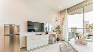 Una habitación de hotel moderna con una cama, un televisor de pantalla plana, un refrigerador, un tocador blanco, un espejo redondo, una planta en maceta y un balcón con vistas a la ciudad.