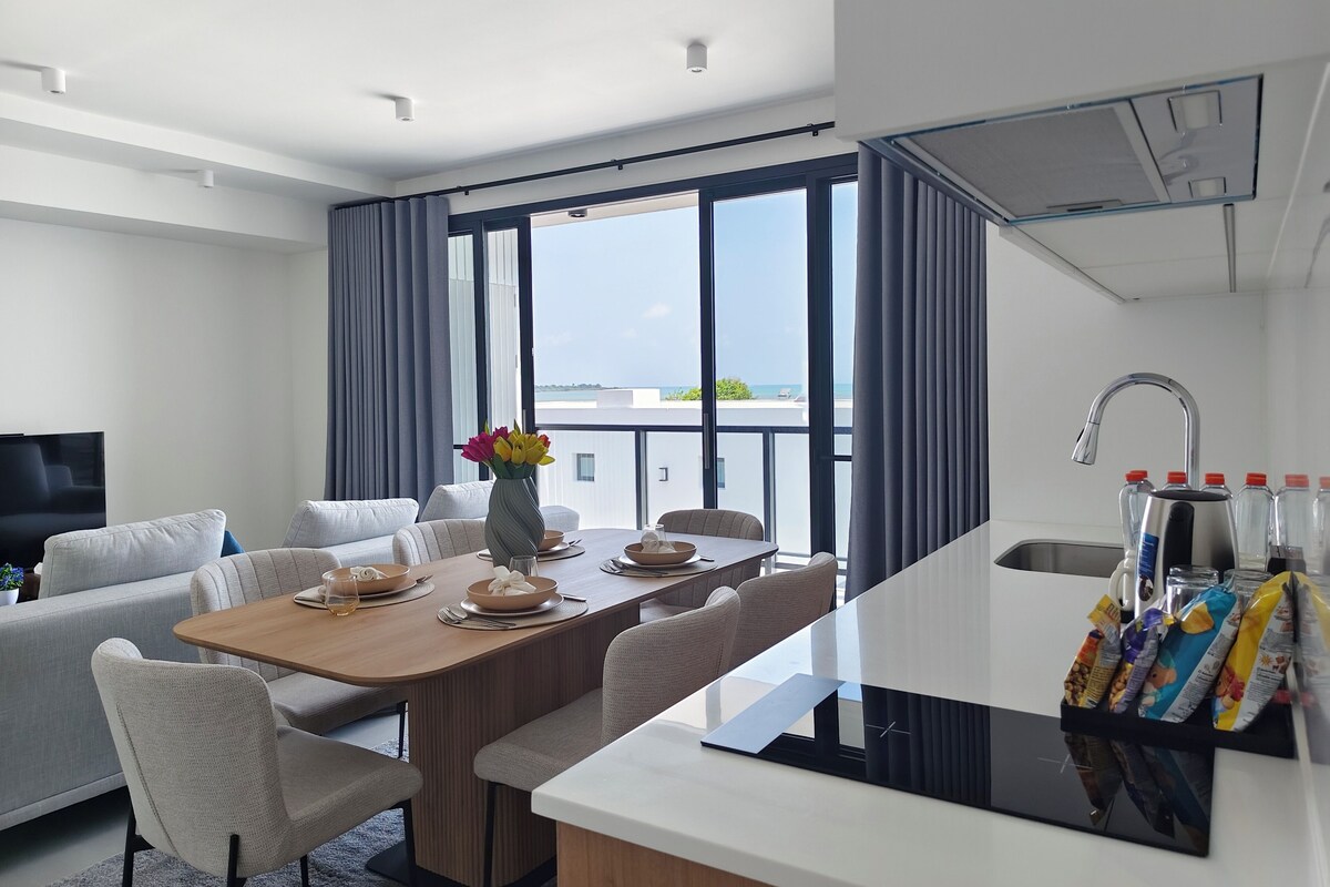 A modern hotel room with a dining area, a sofa, a TV, and a view of the sea.