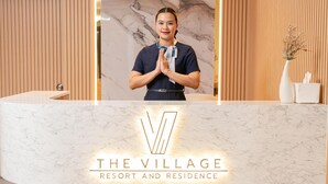 A reception desk with a marble top, a logo reading 'THE VILLAGE RESORT AND RESIDENCE,' and a person standing behind it. - The Village Chumphon Hotel (Pathio)