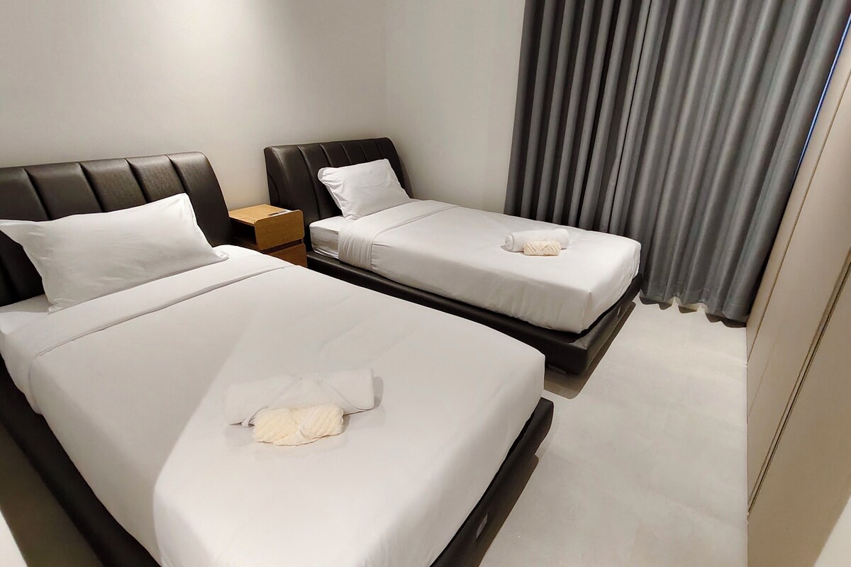 Habitación de hotel con dos camas, una mesita de noche de madera y una ventana con cortinas grises.