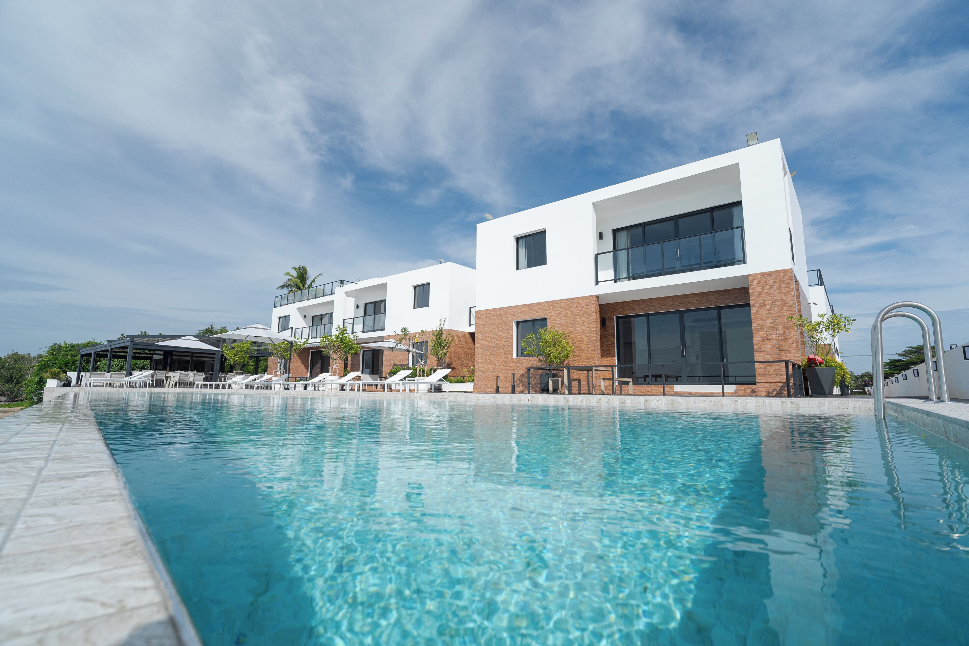 Un área de piscina moderna con agua azul cristalina, sillones de descanso y un edificio de fachada de ladrillo y amplias ventanas.