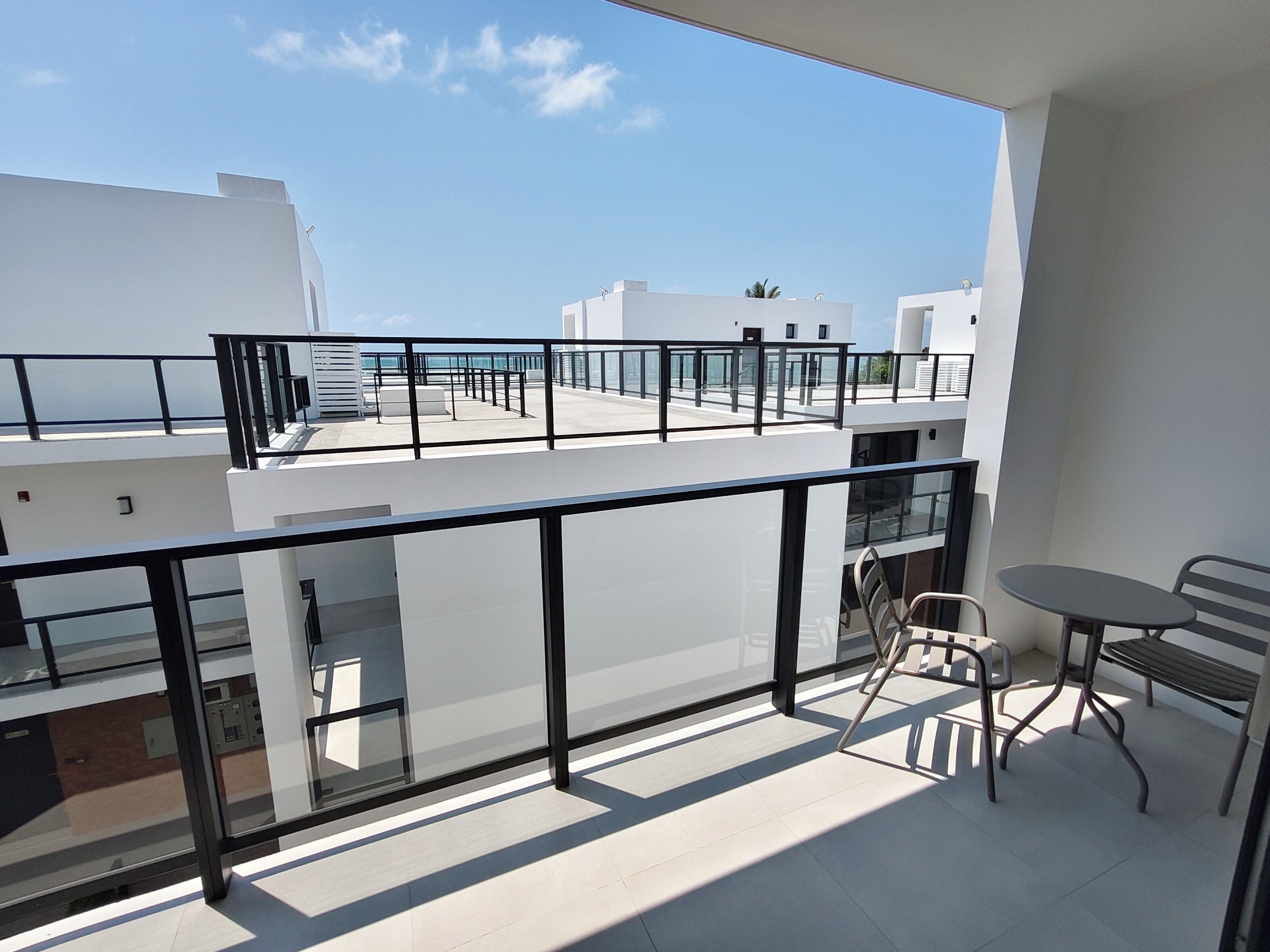 A balcony with a glass railing, a table, and a chair, overlooking a clear sky and buildings.