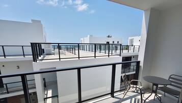 A balcony with a glass railing, a table, and a chair, overlooking a clear sky and buildings.