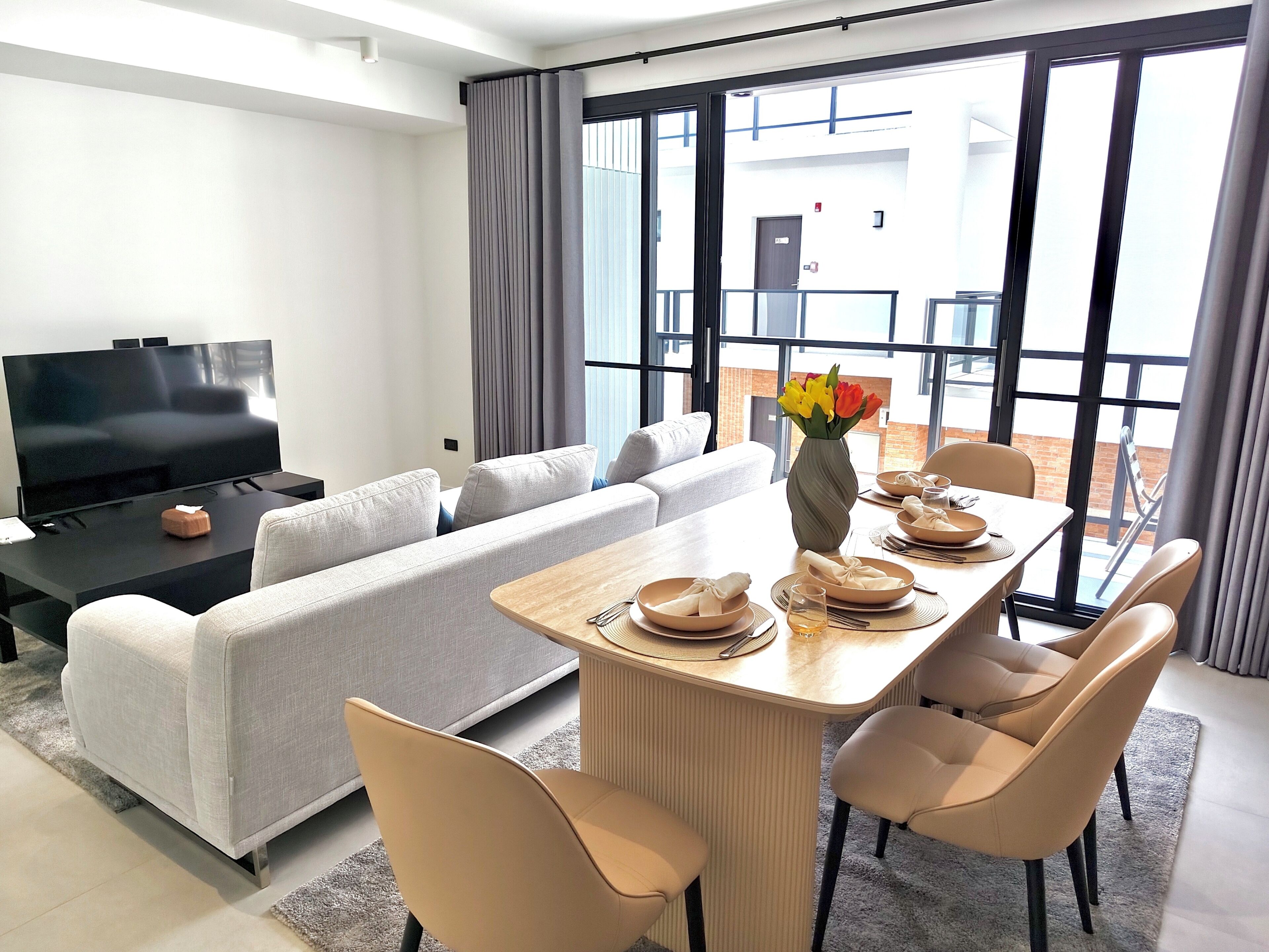 Una sala de estar moderna con televisor de pantalla plana, sofá gris, mesa de comedor para cuatro personas y un amplio ventanal con vista al exterior del edificio.