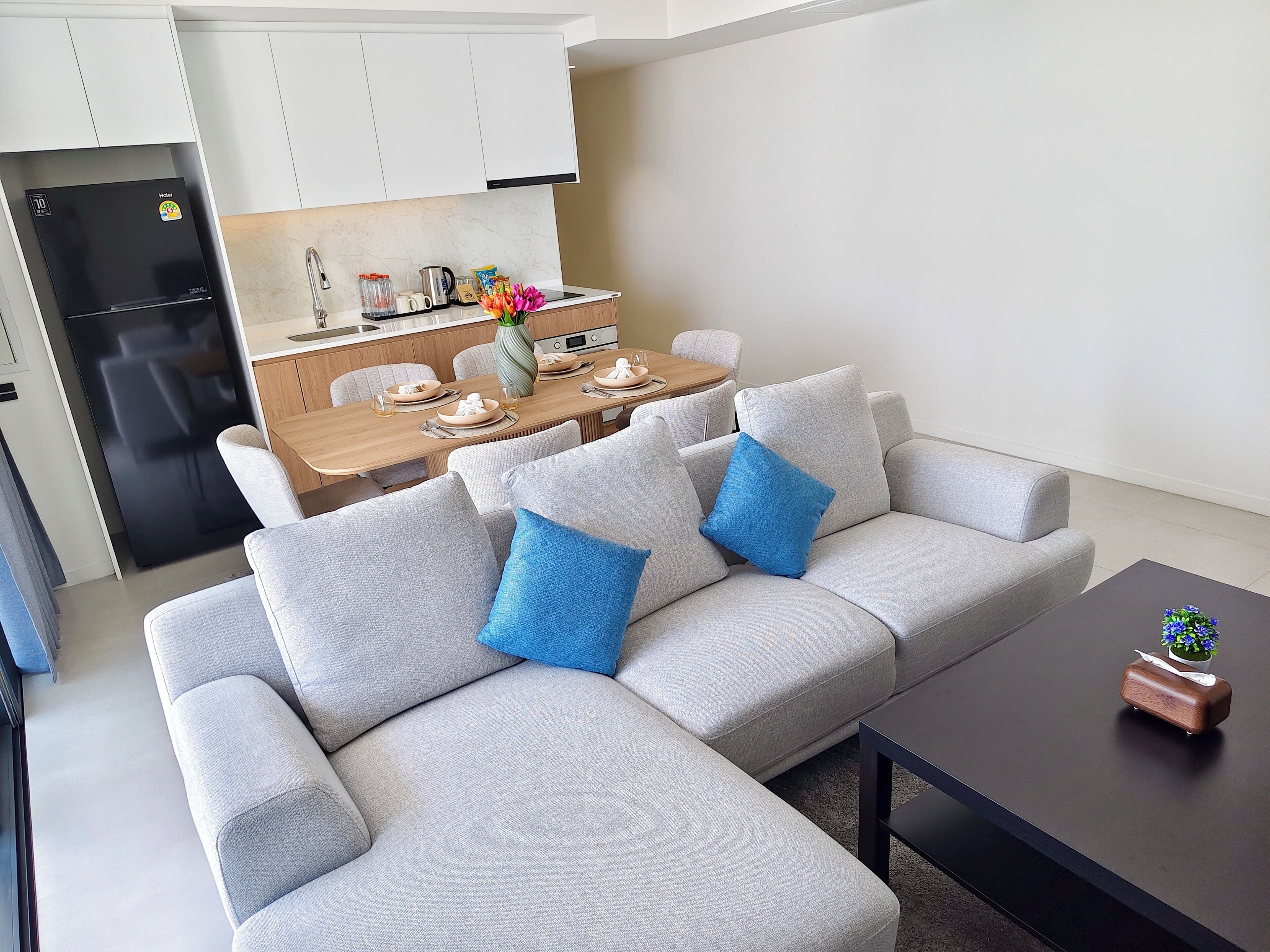 Una sala moderna con un sofá seccional gris, una mesa de comedor para cuatro personas, un refrigerador negro y una pequeña mesa con un objeto decorativo.