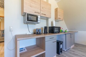 Una kitchenette con microondas, tostadora y cafetera sobre la encimera, un cubo de basura y armarios de madera.