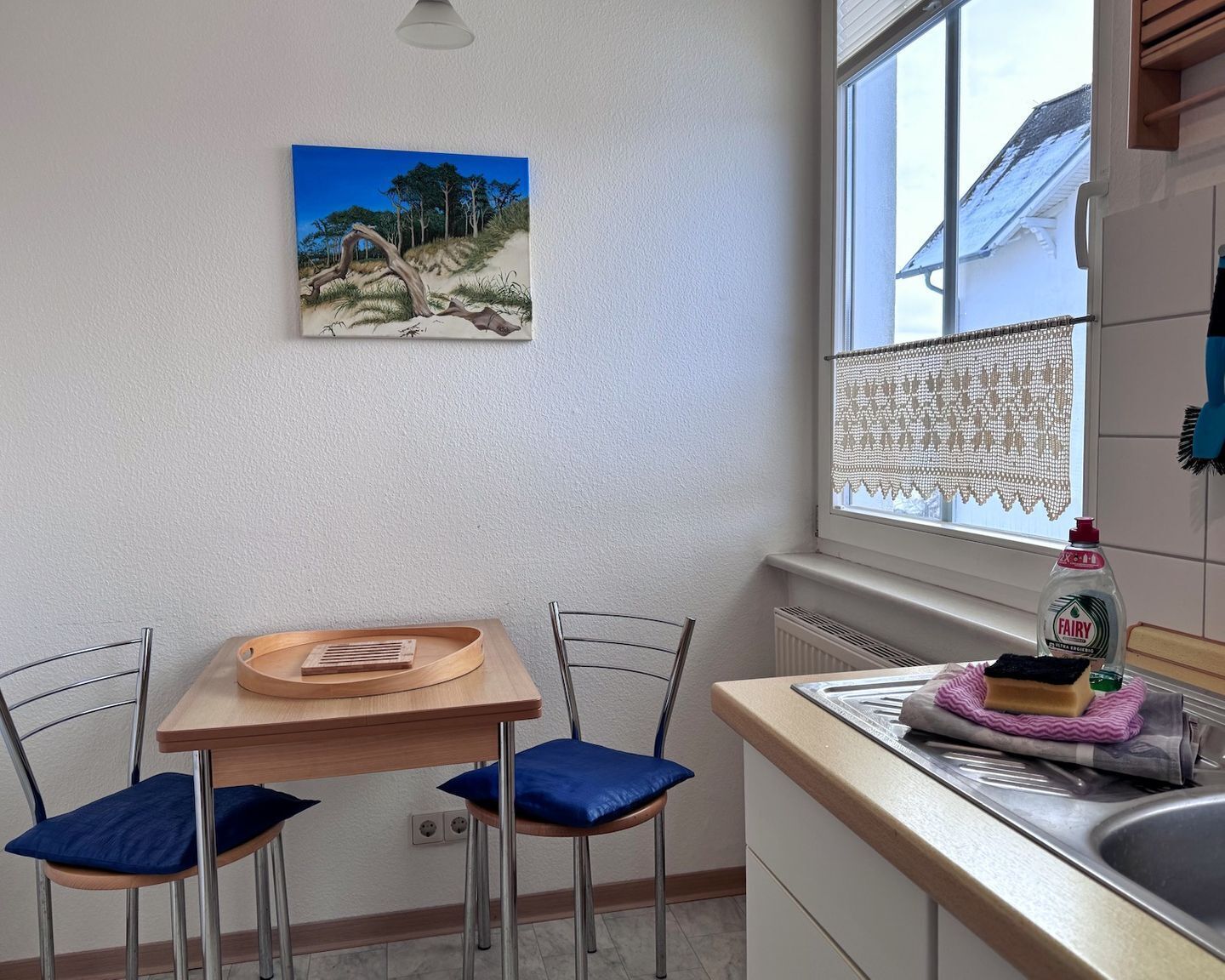Eine kleine Küche mit einem Holztisch, zwei Stühlen, einem Bild an der Wand, einem Fenster mit Spitzenvorhängen und einer Flasche Fairy-Spülmittel auf der Arbeitsplatte.