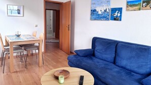 Una sala de estar con un sofá azul, una mesa de comedor de madera con sillas, y una mesita redonda con un bol y un control remoto.