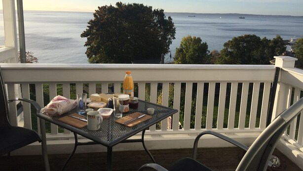 Un balcón con una mesa dispuesta para el desayuno, con vista a una masa de agua y barcos a lo lejos.
