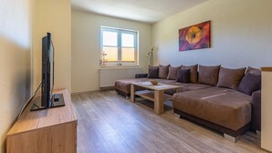 A living room with a sofa, a coffee table, a TV on a stand, and a window with blinds. - Pferdehof Königsstuhl - Apartment 5 Friese - Holidays at the riding stable - Apartment (Lohme)