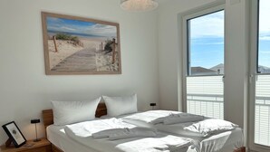 Ein Schlafzimmer mit einem hölzernen Bett, weißer Bettwäsche, einem Nachttisch mit Lampe, einem gerahmten Bild an der Wand und Blick auf den Strand durch das Fenster.
